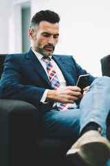 Pensive mature entrepreneur dressed in formal wear reading financial news on website on smartphone...