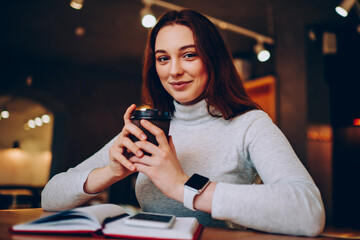 Portrait of positive brunette female student resting on coffee break in cafeteria, smiling young woman in casual wear enjoying aroma beverage for starting creation of article in notebook for blog