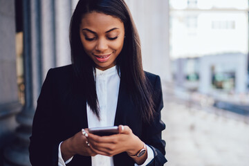 Positive african american businesswoman dressed in formal wear laughing during messaging online on smartphone device via 4G internet.Cheerful dark skinned female office worker installing application