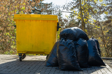 Colorful yellow trash container sits next to a heap of black garbage bags, surrounded by trees and greenery in a peaceful urban park under the warm sunlight