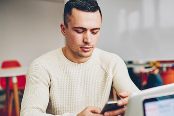 Serious businessman reading financial news using application on smartphone sitting in coworking space, concentrated male entrepreneur browsing web page on mobile phone for making banking online