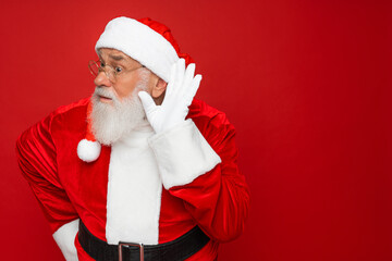 Old bearded Santa Claus man wears Christmas hat red suit try hear you overhear listening intently isolated on plain red background studio. Happy New Year 2025 celebration merry ho x-mas concept