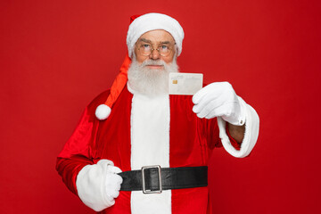 Bearded Santa Claus in Christmas hat red coat holding in hand using showing demonstrate plastic bank credit card store shop cash back cashback isolated bright vivid shine vibrant red color background