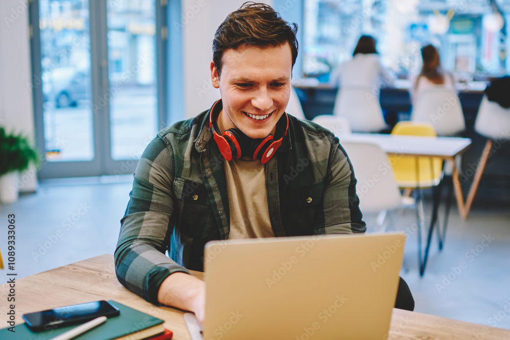Poster cheerful hipster guy with headphones enjoying playing computer games spending leisure with laptop in
