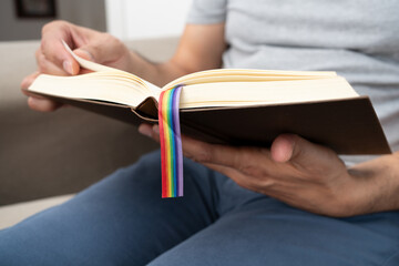 Happy man reading a paper book with LGTB flag sitting on a sofa at home