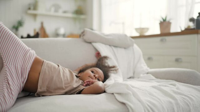 Funny little African girl lying on soft couch in living room, lazy weekend. Charming four-year-old child relaxing on sofa at home, comfortable and cozy apartment, carefree childhood, mixed race family