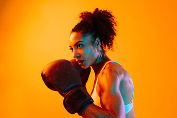 Sportive black hispanic woman wearing sportswear training boxing in fitness studio, colorful dramatic lighting - African-american female athlete doing sport workout