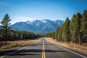 Fototapeta premium Empty stretch of asphalt road running parallel to a mountain range with a clear blue sky above and pine trees in the foreground, mountain view, natural landscape, empty highway
