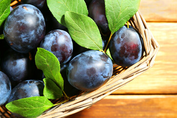 Tasty ripe plums and leaves in wicker basket on wooden table, top view