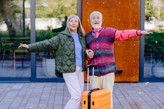 Elderly man and woman standing outside modern building with orange suitcase. Both are extending arms like airplane, wear cozy, colorful jackets and appear relaxed, spirit of travel and adventure. - Powered by Adobe