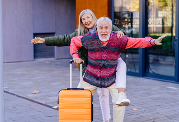 Elderly couple enjoying travel together outside modern building. Senior man and woman express happiness and freedom, posing playfully with bright orange suitcase. Adventure, love, active lifestyle.
