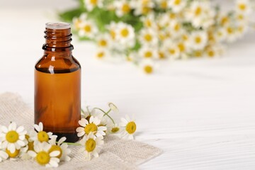 Bottle of essential oil and chamomile flowers on white wooden table, closeup. Space for text