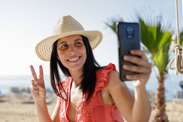 Young woman taking a selfie with her cell phone to send to her friends, family and social networks. Young woman with her straw hat smiles on vacation enjoying a sunny day.