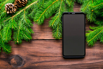 A black smartphone with a bright black screen lies on a wooden surface, surrounded by festive fir branches and pinecones, creating a holiday-themed aesthetic.

