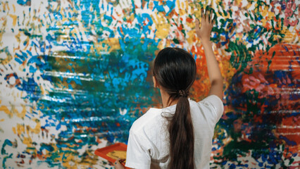 Obraz premium Young happy girl holding the color tray and paint colorful stained wall to express idea and enhance imagination with hand print. Caucasian girl stamp hand to decorate room in art lesson. Edification.