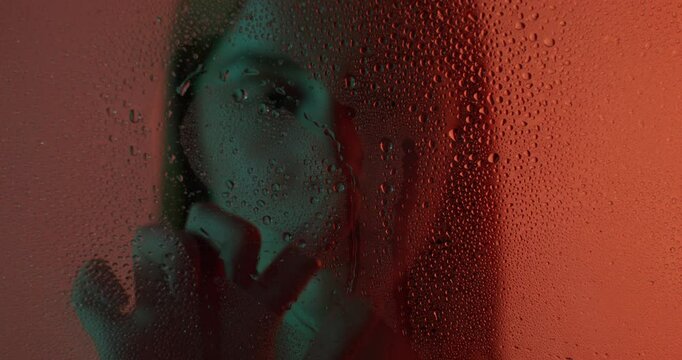 A woman draws a heart on the glass of a shower stall with drops of water. She looks at the camera with a serious look. View through the glass