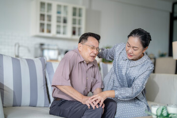 An elderly couple sits in their living room. The man holds his knee, appearing to be in discomfort, while his partner offers support. The image conveys care, concern, and the bond between the couple.