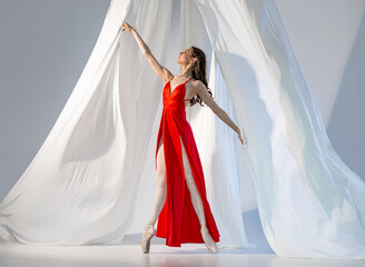 Ballerina in red long dress dancing on pointe shoes in white light studio with white cloth