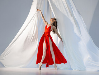 Ballerina in red long dress dancing on pointe shoes in white light studio with white cloth