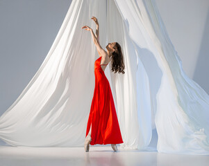 Ballerina in red long dress dancing on pointe shoes in white light studio with white cloth