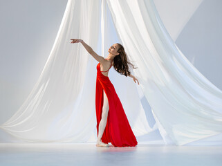 Ballerina in red long dress dancing on pointe shoes in white light studio with white cloth
