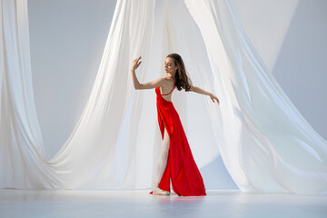 Ballerina in red long dress dancing on pointe shoes in white light studio with white cloth