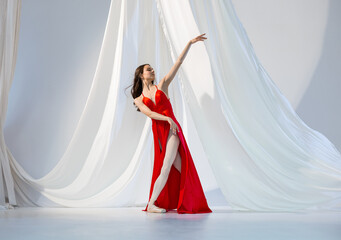 Ballerina in red long dress dancing on pointe shoes in white light studio with white cloth
