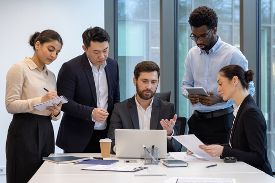 Professional business team engaged in collaborative meeting around laptop, discussing strategies. Group includes diverse individuals with documents, notepad, and tablet, enhancing teamwork