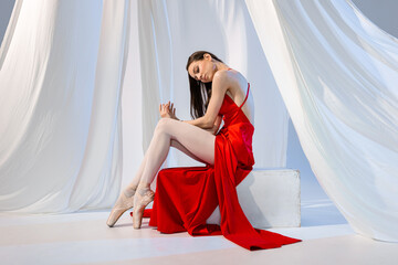 Ballerina in pointe shoes in red long dress sitting on the white cube in white light studio 