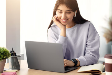 People And Modern Technology Concept. Portrait of smiling woman in wireless headphones sitting at desk, using laptop and watching tutorial, lecture or webinar, studying online at home