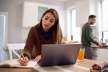 Mid adult woman taking notes while working on laptop at home.