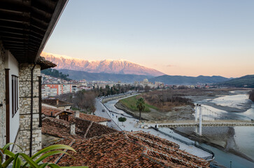 A panoramic view of a Berat with the Osumi river flowing through it. The city is nestled against a backdrop of snow-capped mountains, with a traditional bridge spanning the river. 