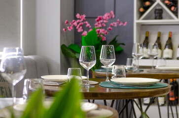 A cozy restaurant interior with round wooden tables set with white plates and wine glasses. The tables are decorated with green plants and pink flowers. A wine rack with bottles is visible in the back