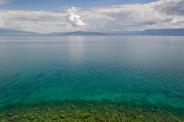  A serene Ohrid lake stretches out towards a distant mountain range in North Macedonia under a bright blue sky with fluffy white clouds. The crystal-clear water reveals a shallow, rocky bottom.