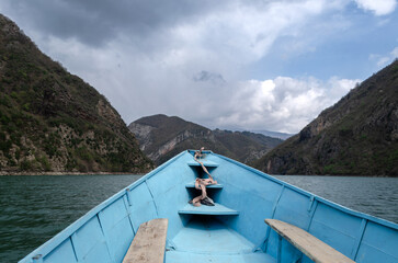 A small, blue boat glides through a serene lake surrounded by towering, lush mountains. The sky is filled with dramatic, fluffy clouds, casting a serene atmosphere.