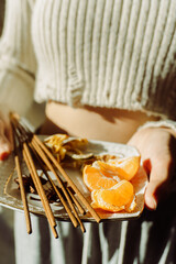 Hands holding a tray with cinnamon sticks, orange slices, and dried fruit, illuminated by soft sunlight, creating a warm and cozy atmosphere