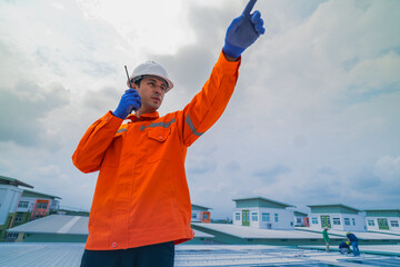 A worker in an orange safety jacket inspects solar panels on a rooftop, using a laptop to ensure...