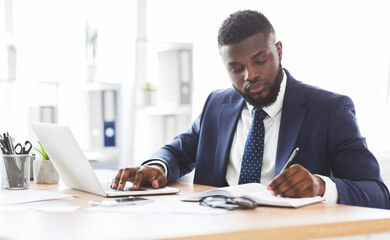 Successful american businessman working hard in office, making notes in front of laptop, panorama...