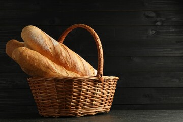 Fresh baguettes in wicker basket on black table, space for text