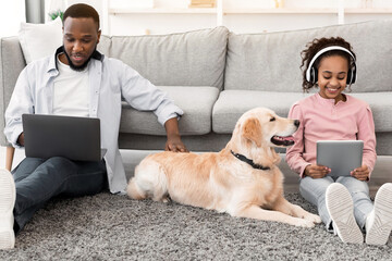 Leisure Time. African American girl sitting on rug floor carpet with dog and holding digital...