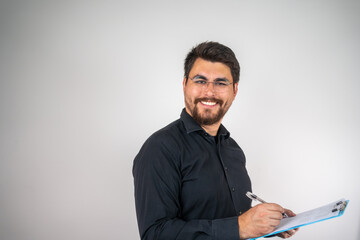 Young man with beard, glasses writing in clipboard standing isolated on grey background.
