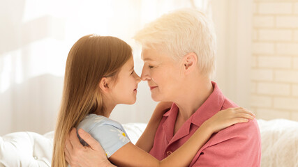 Family happiness concept. Lovely little girl and her grandma touching noses and embracing at home