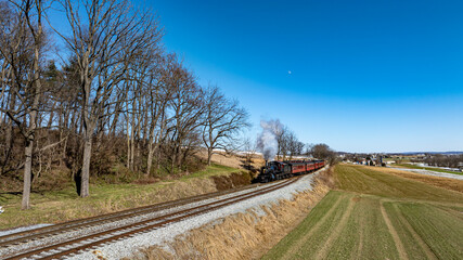 A historic steam locomotive chugs along the tracks, billowing smoke against a clear blue sky while surrounded by lush green fields and distant hills