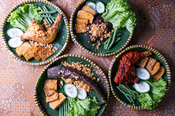 An overhead view of a vibrant and appetizing spread of Nasi Ayam Penyet served on banana leaves, surrounded by fresh vegetables, crispy fried toppings, and sambal-coated chicken.