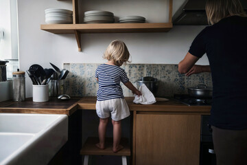 Child standing on a stool helping parent in a cozy modern kitchen with wooden cabinetry and shelves filled with plates
