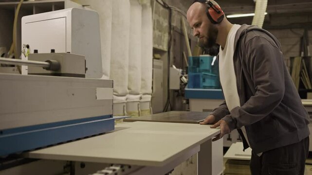 Professional carpenter in protective earmuffs operating edge banding machine on wood panel in furniture workshop