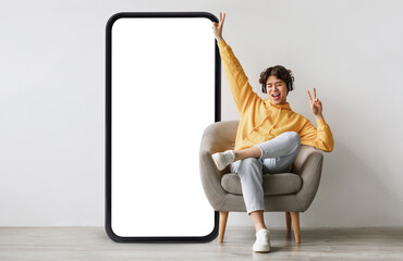 Joyful guy sittiwng on chair near big empty smartphone screen, listening to music in wireless...