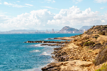 A stunning coastal scene featuring rugged rocky shores, turquoise waters, and a distant city skyline framed by mountains. Alicante, Spain. 