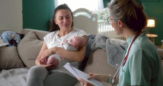 Female doctor and patient mother with newborn baby discussing test results looking at papers and talking in living room at home. 