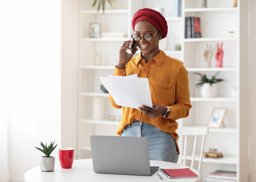 Young businesswoman cheerful african american lady with red turban on her head working at office, standing by workdesk with laptop, holding documents, having phone conversation, copy space - Powered by Adobe